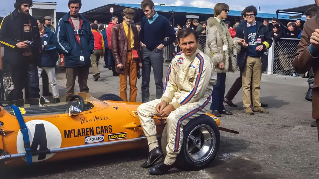بروس مکلارن (Bruce McLaren) در کنار خودروی مسابقه‌ای نارنجی رنگ مک‌لارن (McLaren)
