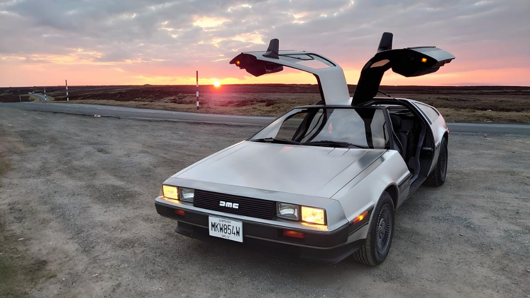 خودرو دیلورین (DeLorean) با درهای بال‌پرنده در مسیر جاده‌ای هنگام غروب
