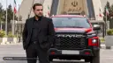مردی در کنار خودروی قرمز برند کرمان موتور (KMC) در مقابل ساختمان مجلس