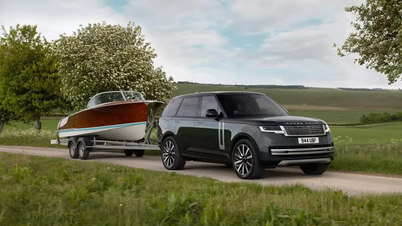 رنجرور الکتریک (Range Rover Electric) در حال حرکت و یدک‌کشی یک قایک کلاسیک با تریلر در مسیر روستایی